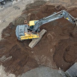 KERBUD - Żółto-szara koparka Volvo na placu budowy z ziemią i kostką brukową, widziana z góry. Maszyna w trakcie pracy, z łyżką skierowaną w dół. Widoczne logo firmy na maszynie.