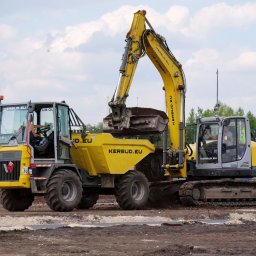 KERBUD - Żółto-szara koparka gąsienicowa z łyżką wypełnioną ziemią, obok wywrotka na placu budowy z widocznymi operatorami w kabinach i przygotowanym podłożem.