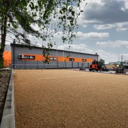 Przygotowany teren pod budowę parkingu lub placu, podsypka z kruszywa, obrzeża betonowe, w tle budynek z pomarańczowymi pasami, maszyny budowlane i pracownicy w kamizelkach odblaskowych.