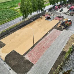 Widok z góry na plac budowy z częściowo ułożoną kostką brukową w odcieniach szarości i czerwieni, przygotowany teren pod kostkę z piaskową podsypką, maszyny budowlane w tle i palety z kostką,...