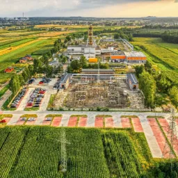 Szeroki widok z lotu ptaka na teren przemysłowy z budynkami, parkingiem, transformatorami i polem kukurydzy w słoneczny dzień.
