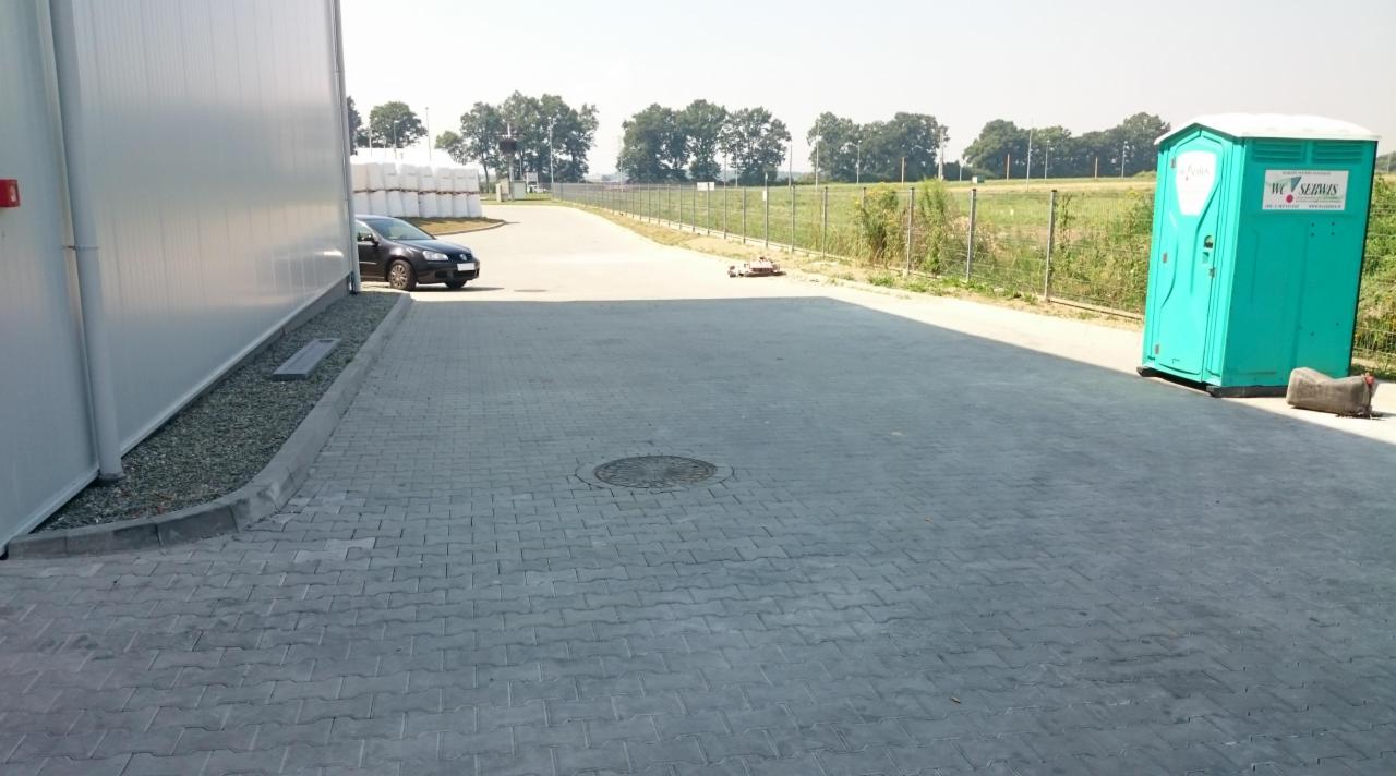 Szara kostka brukowa na placu przemysłowym, widoczny budynek magazynowy, zaparkowany samochód, przenośna toaleta WC Serwis oraz oddalona zieleń i drzewa.