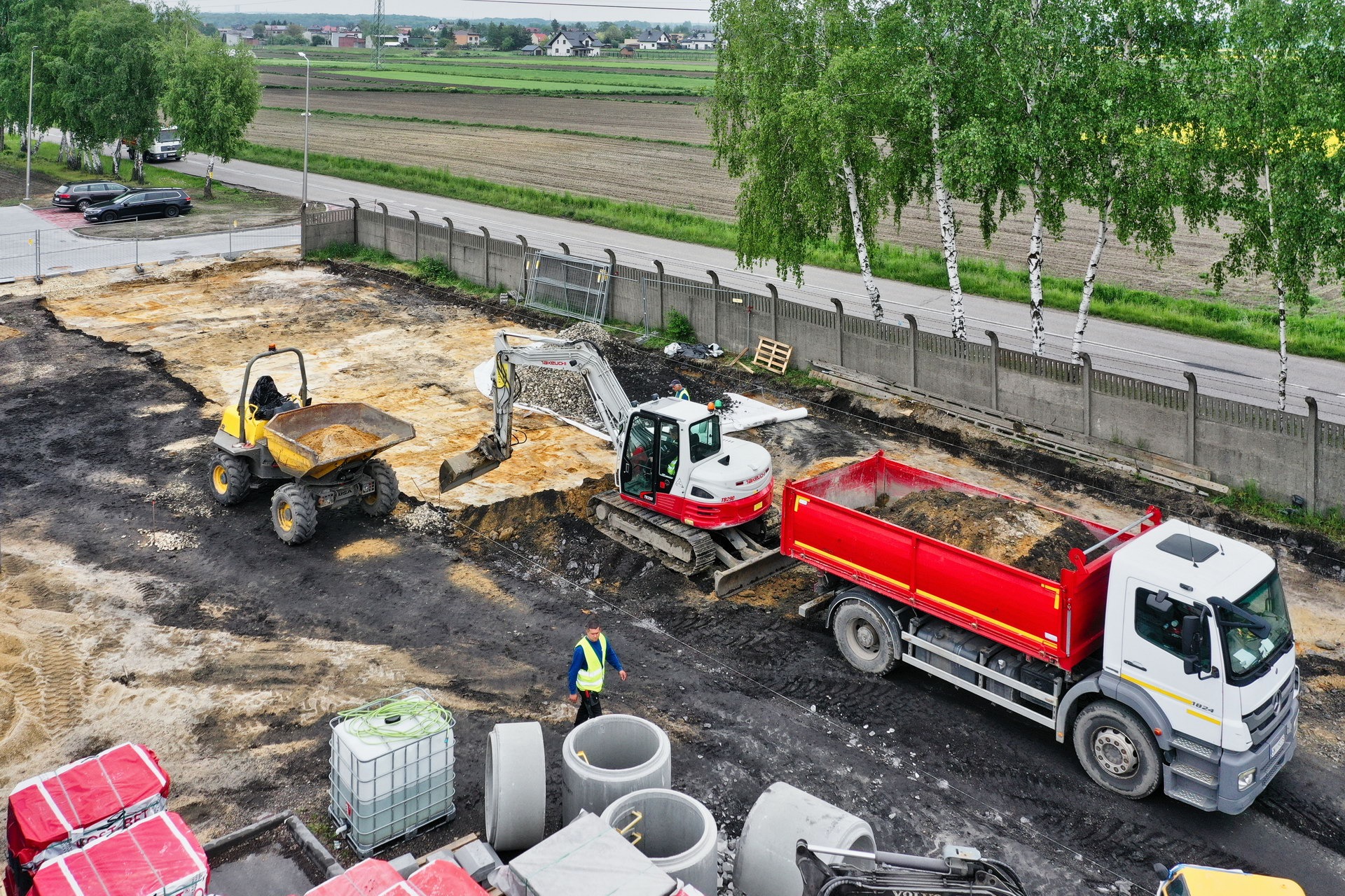 Widok z góry na plac budowy z koparką, wywrotką, dumperem, pracownikiem w kamizelce odblaskowej, betonowymi rurami i workami z materiałem, teren ogrodzony betonowym płotem z drutem kolczastym.