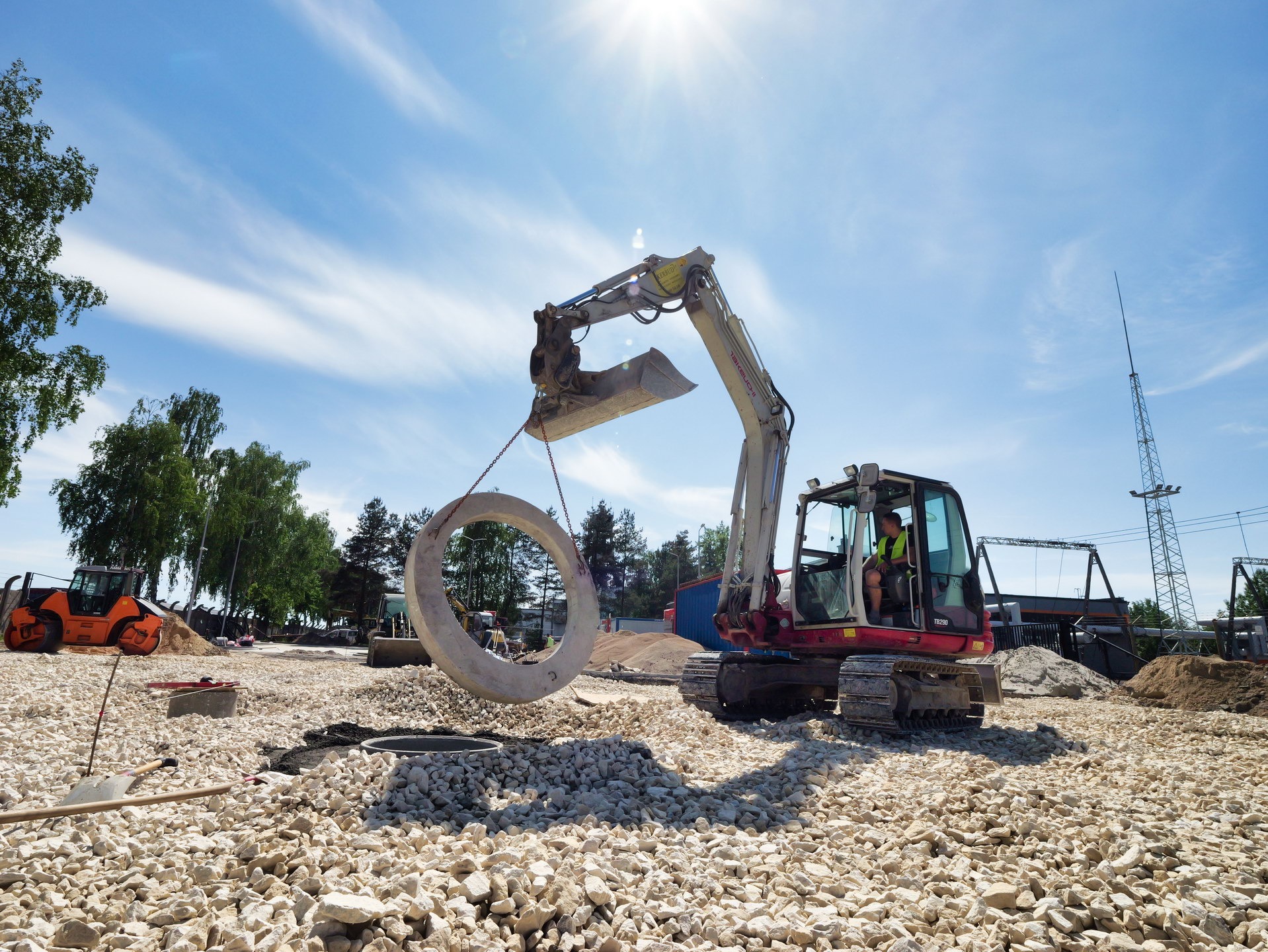Czerwono-biała mini koparka podnosi betonowy pierścień na placu budowy z utwardzonym podłożem z białego kruszywa, w tle widoczny walec drogowy i słupy energetyczne.