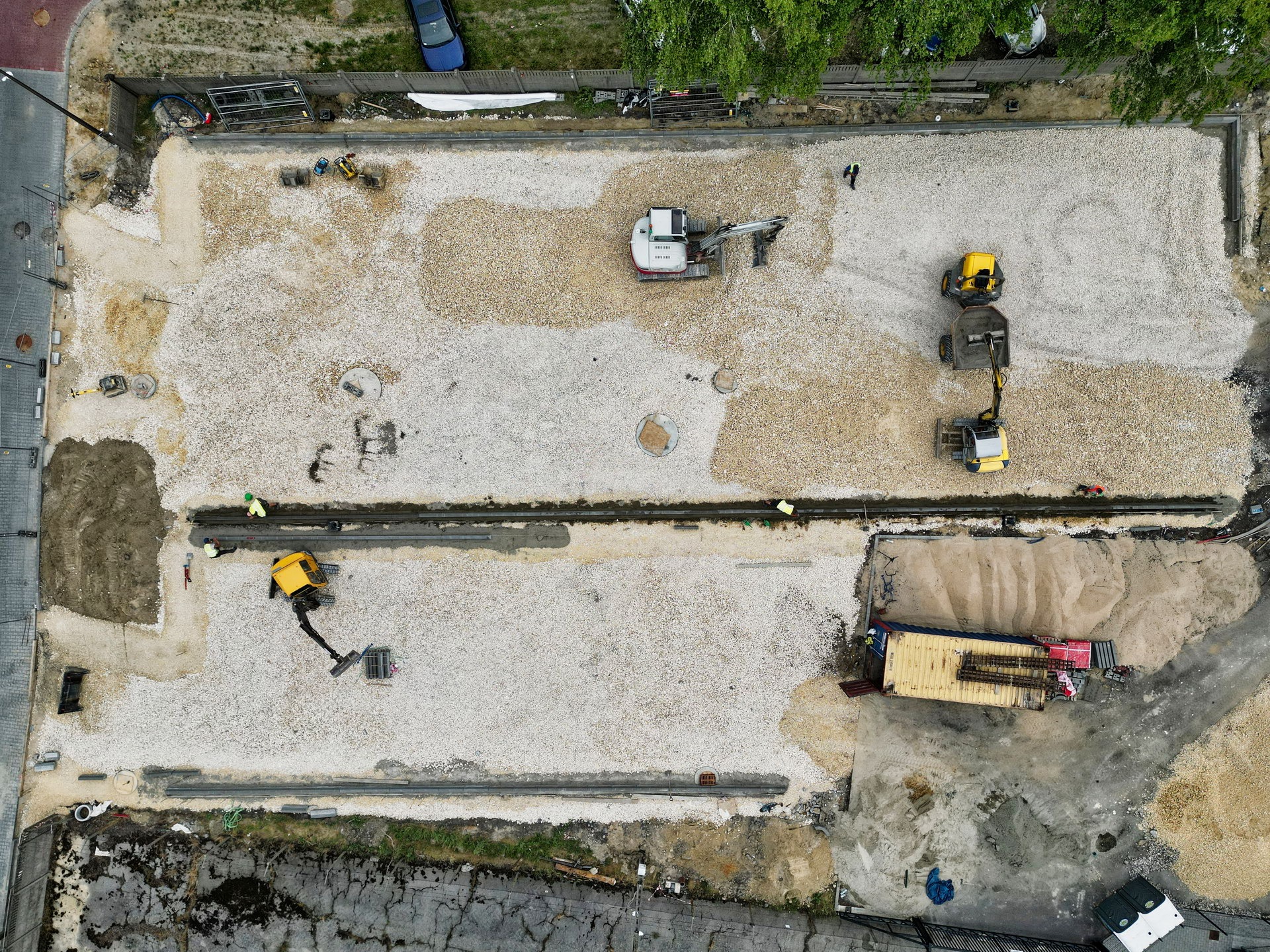 Widok z góry na plac budowy z rozłożonym jasnym żwirem, kilka maszyn budowlanych w trakcie prac ziemnych, widoczne fundamenty pod przyszłą konstrukcję.