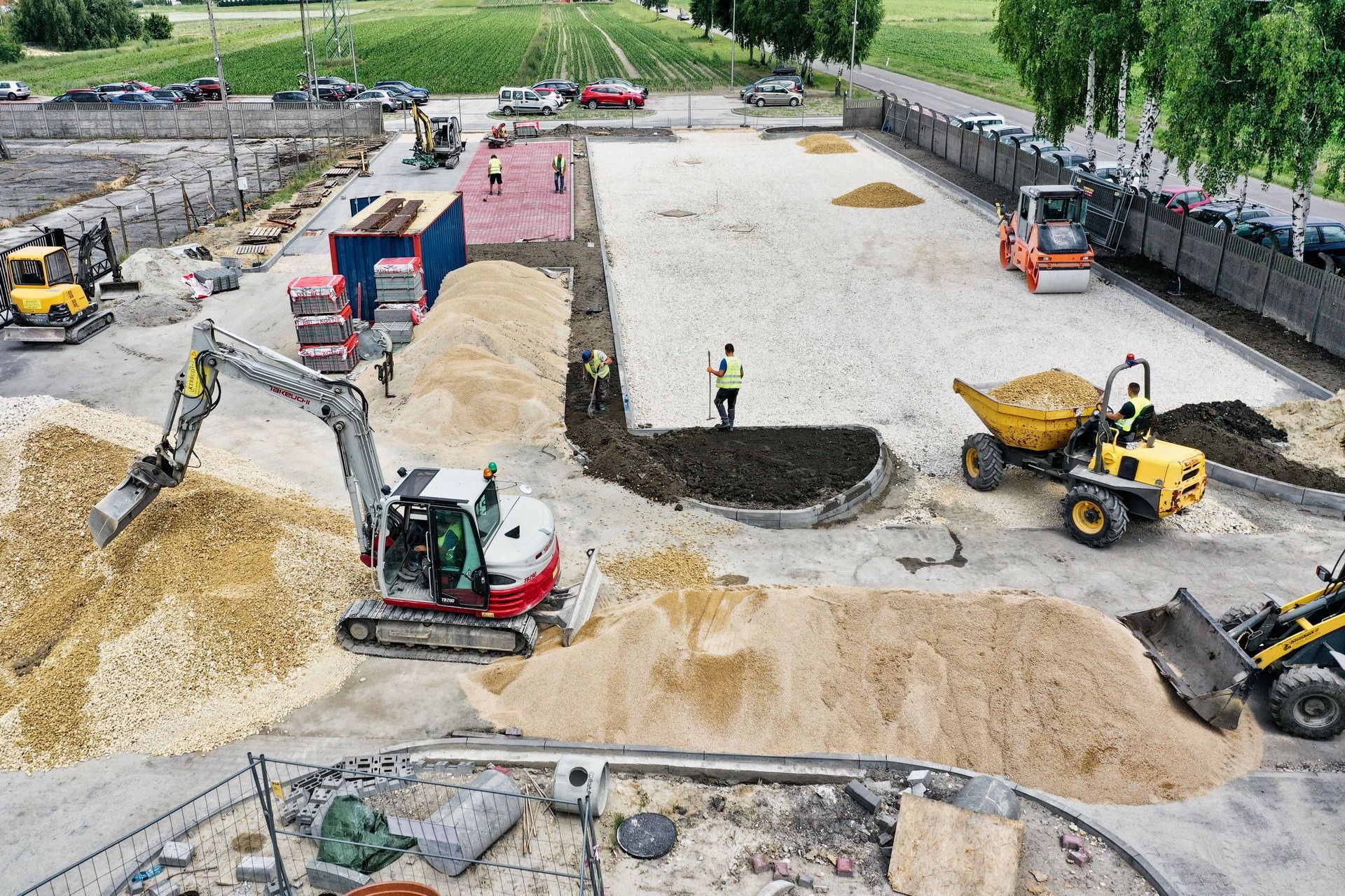 Widok z góry na plac budowy z maszynami: koparka z łyżką nabierająca piasek, walec drogowy, wywrotka z piaskiem, robotnicy w kamizelkach odblaskowych przy pracach ziemnych. W tle zaparkowane...
