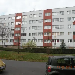 Wielopiętrowy budynek z szarą elewacją i czerwonymi akcentami, widok z ulicy z zaparkowanymi autami i zielenią. Widoczne anteny satelitarne na ścianie.