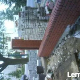 Murowanie z cegły klinkierowej, widoczny fragment muru z cegły, stos cegieł i wiadro, etap budowy ogrodzenia.