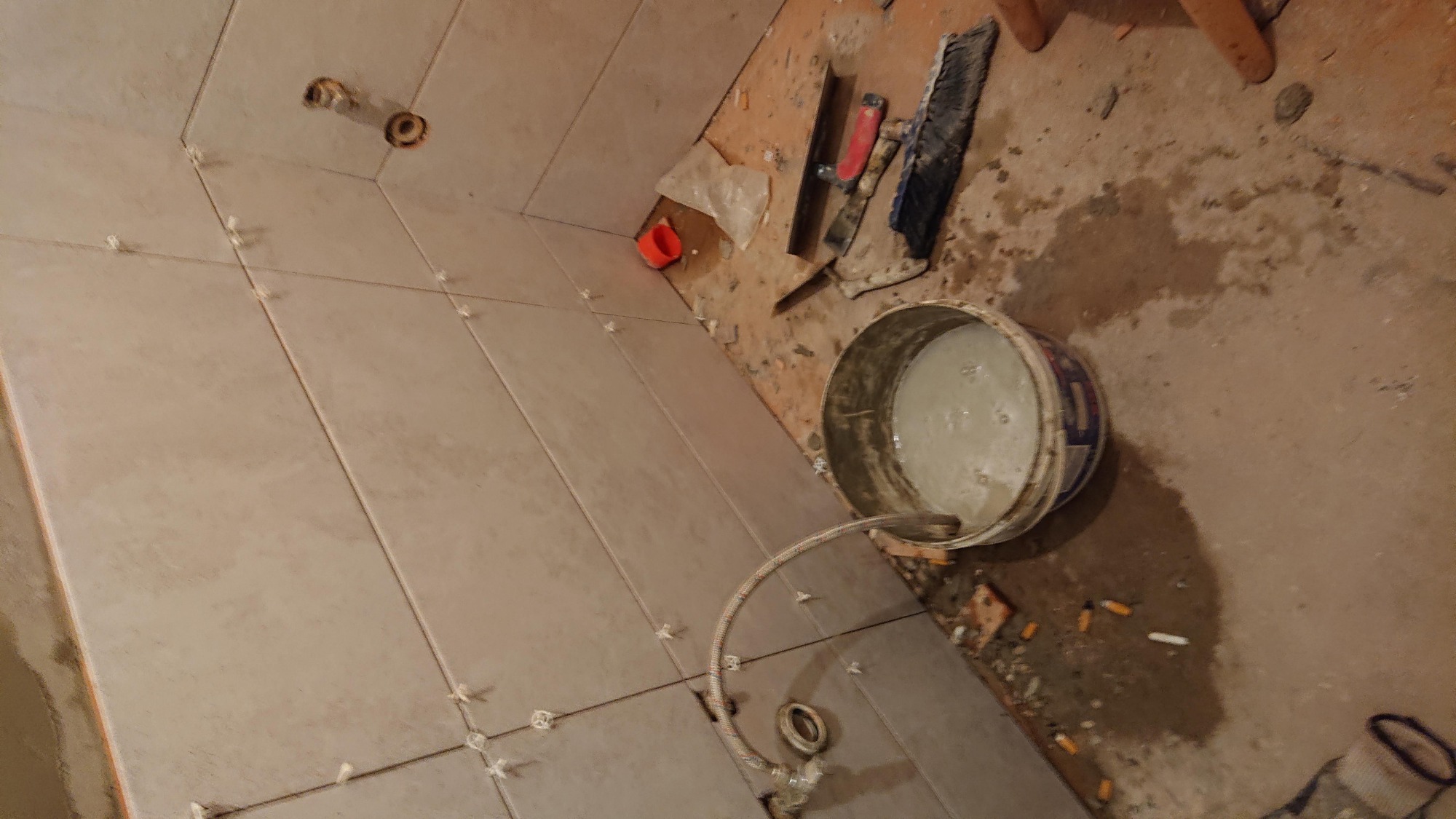 Układanie płytek w łazience: widok na świeżo ułożone płytki z krzyżykami dystansowymi, wiadro z zaprawą, narzędzia i fragment podłogi w trakcie prac wykończeniowych.