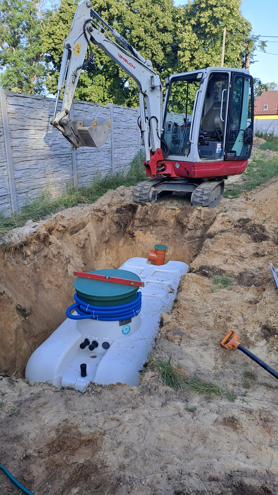 Biały zbiornik z zieloną pokrywą w wykopie, poziomica na górze, niebieskie rury, pomarańczowe elementy. Koparka na górze, szpadel obok. Montaż szamba betonowego.