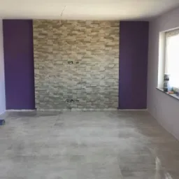 Pokój po remoncie: szare płytki podłogowe, ściany fioletowe i szare. Na środku ściana z kamienia dekoracyjnego. Elementy wykończeniowe przy drzwiach owinięte folią.