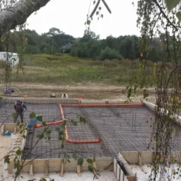 Zbrojenie fundamentu domu jednorodzinnego, widok z góry przez gałęzie drzewa. Na zbrojeniu widoczne pomarańczowe rury. W tle zieleń i biały bus.