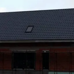 Dom w trakcie budowy z ciemną dachówką i oknem dachowym, otoczony rusztowaniem. Mur z czerwonej cegły, widoczne szare niebo.