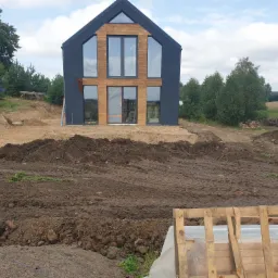 Nowoczesny dom z ciemną elewacją i drewnianymi akcentami, widoczny w trakcie budowy, otoczony ziemią i z żółtą koparką w tle.