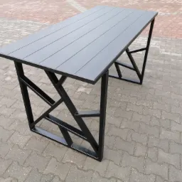 Stół z blatem z desek kompozytowych w odcieniach szarości i nogami z profili stalowych spawanych w geometryczny wzór, stojący na kostce brukowej.
