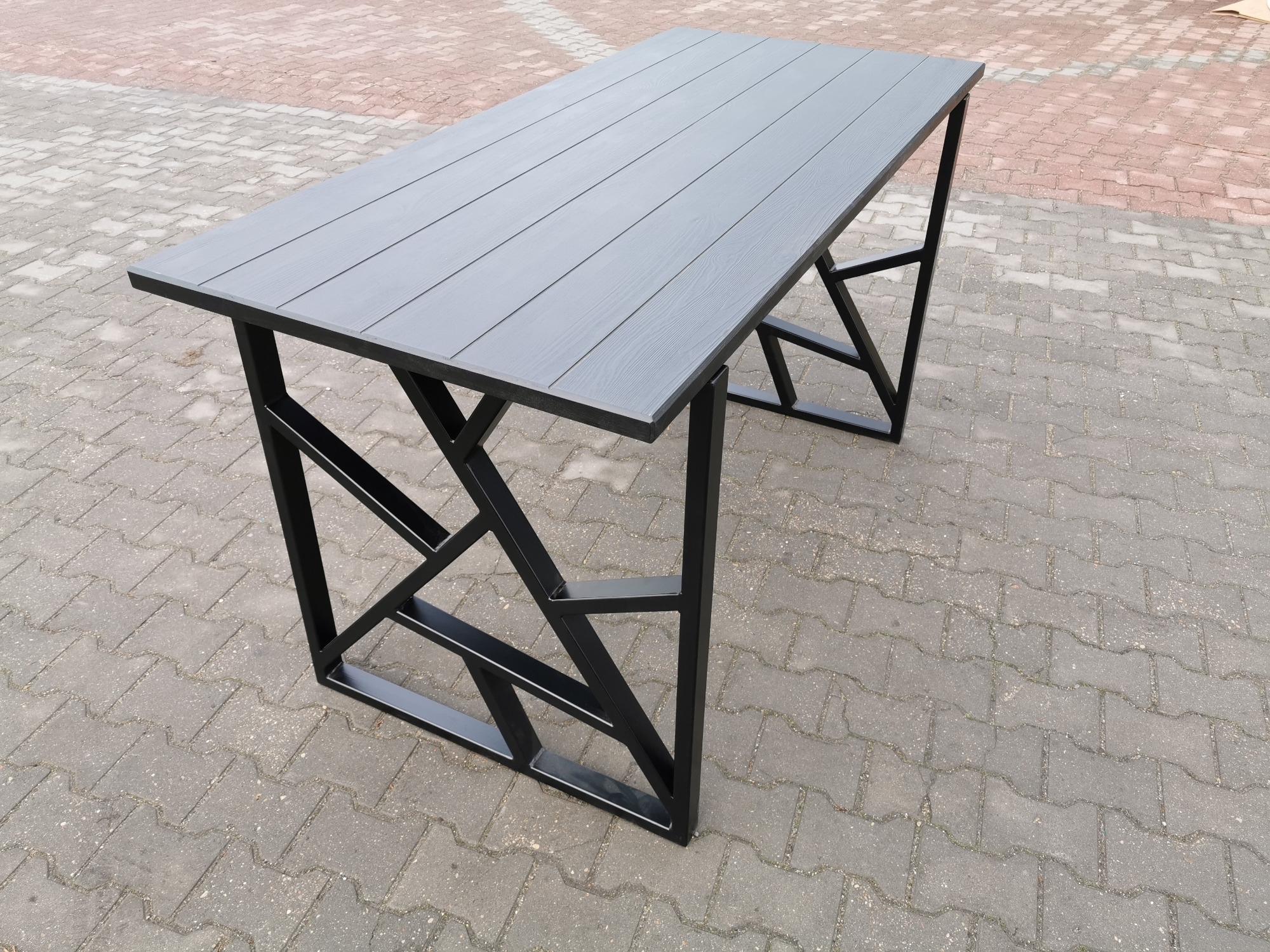 Stół z blatem z desek kompozytowych w odcieniach szarości i nogami z profili stalowych spawanych w geometryczny wzór, stojący na kostce brukowej.