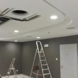 Wnętrze biura w trakcie odświeżania: szare ściany, biały sufit z oświetleniem punktowym i klimatyzacją, dwie aluminiowe drabiny rozstawione w pomieszczeniu, drzwi w tle.