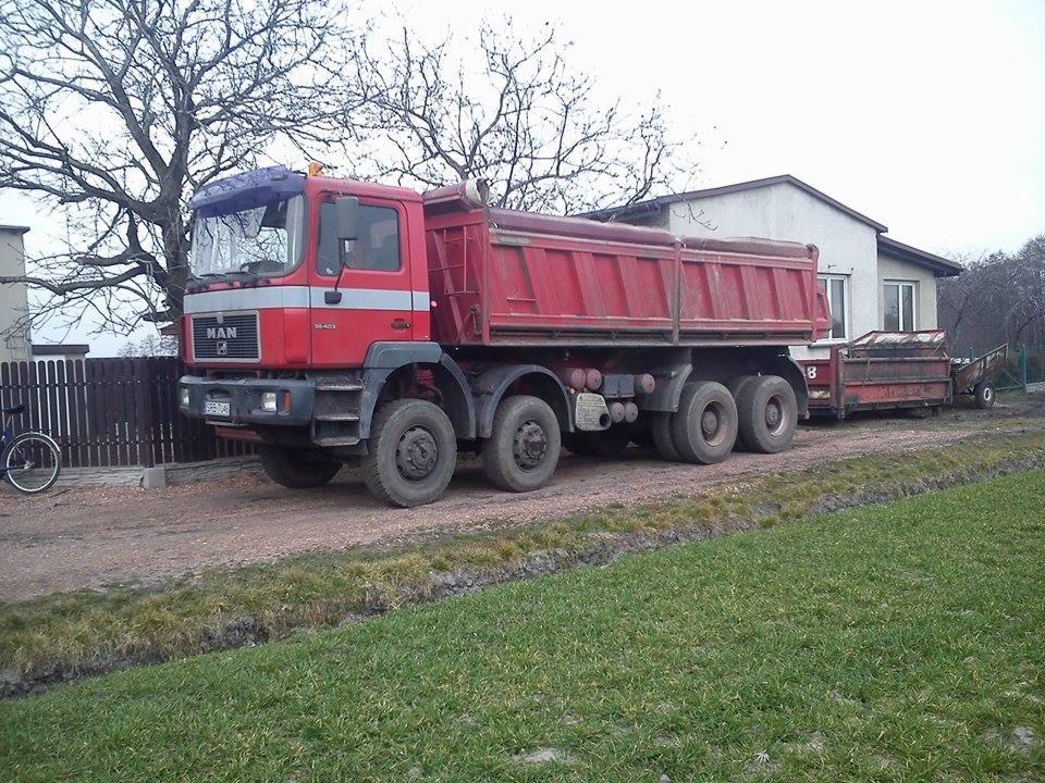 Czerwona ciężarówka MAN z przyczepą na polnej drodze przed domem jednorodzinnym, widok z boku.