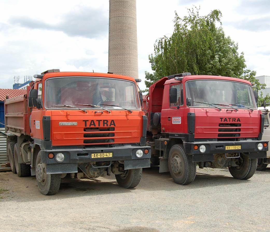 Dwa samochody ciężarowe marki Tatra, jeden pomarańczowy z numerem rejestracyjnym AR-90-47, drugi czerwony z numerem AX 10-42, stoją obok siebie na żwirowym placu, w tle komin i drzewo.