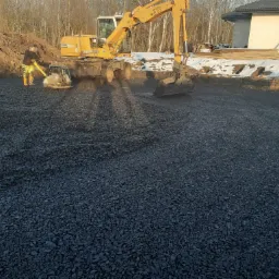 Żółta koparka Liebherr 311 i zagęszczarka wibrująca ubijają czarny żwir na przygotowanym terenie budowy, w tle widoczny częściowo ośnieżony teren i fragment budynku w trakcie budowy.