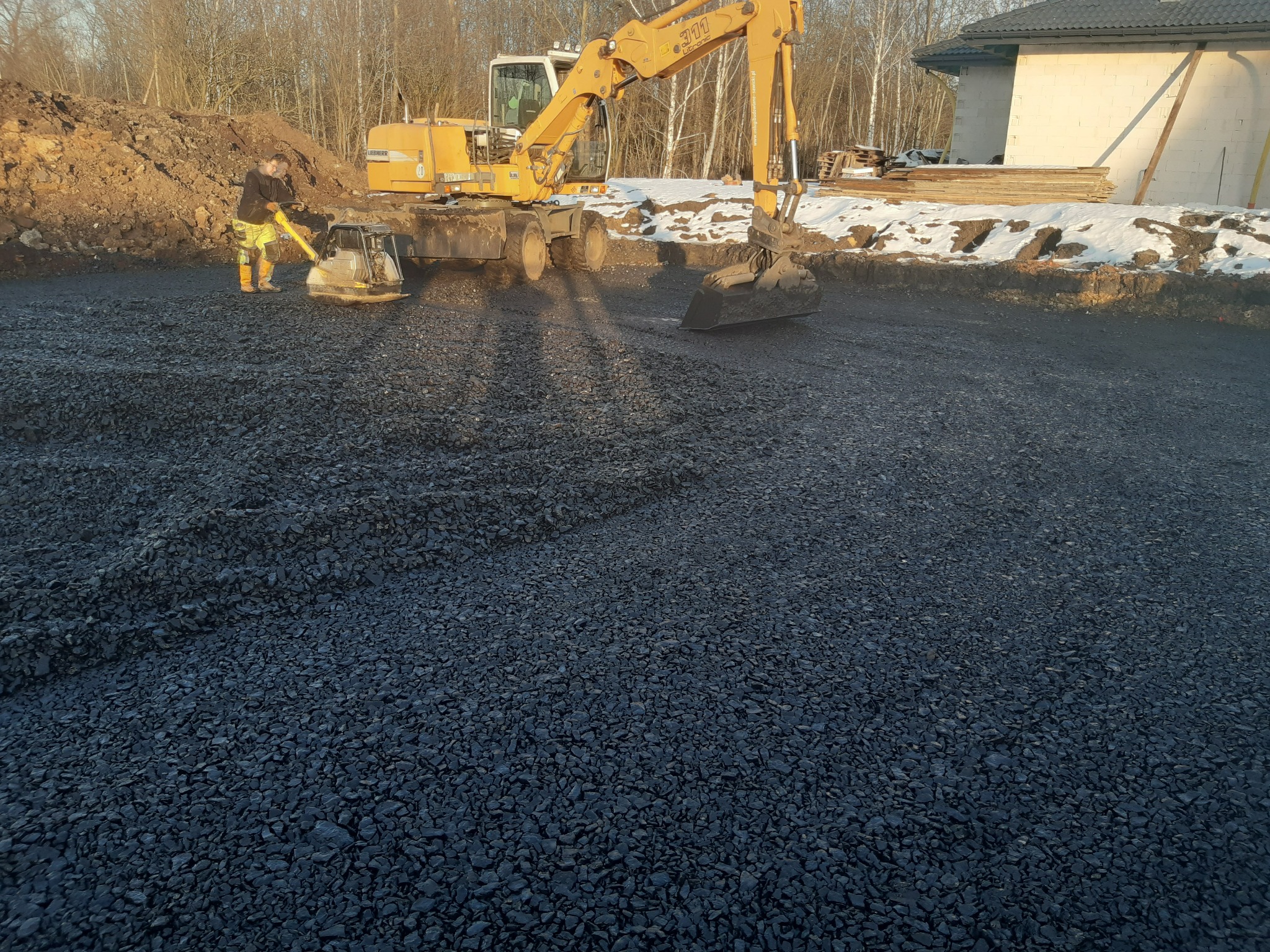 Żółta koparka Liebherr 311 i zagęszczarka wibrująca ubijają czarny żwir na przygotowanym terenie budowy, w tle widoczny częściowo ośnieżony teren i fragment budynku w trakcie budowy.