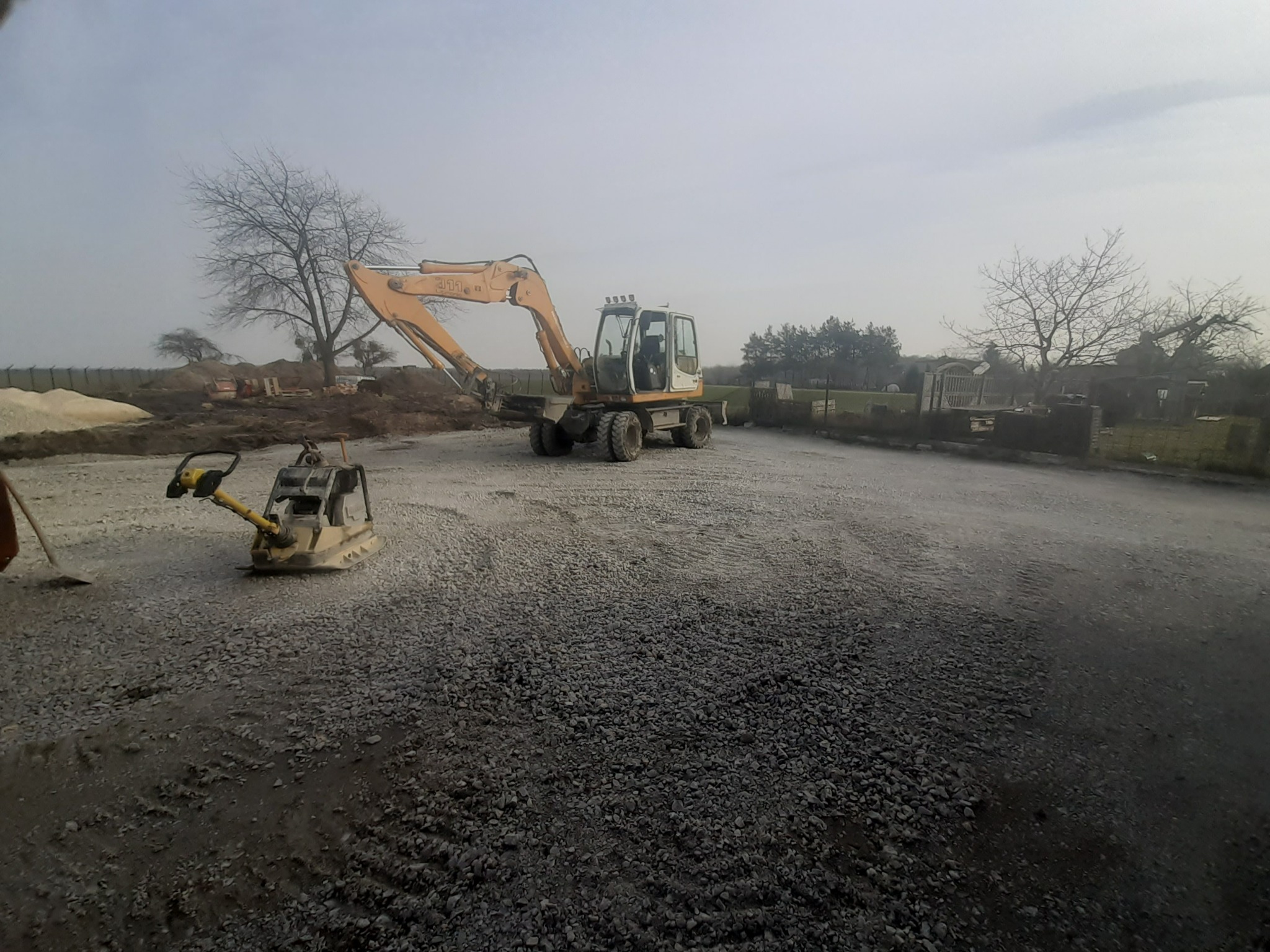 Żółty koparko-ładowarka i zagęszczarka wibrująca na placu budowy, przygotowanym pod fundamenty, w tle drzewa bez liści i mgliste niebo.