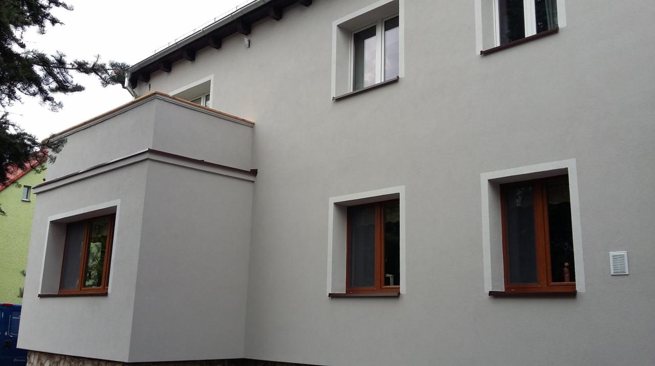 Elewacja budynku po tynkowaniu w odcieniach szarości z brązowymi ramami okiennymi i białymi obramowaniami okien, widok z dołu na fragment budynku.