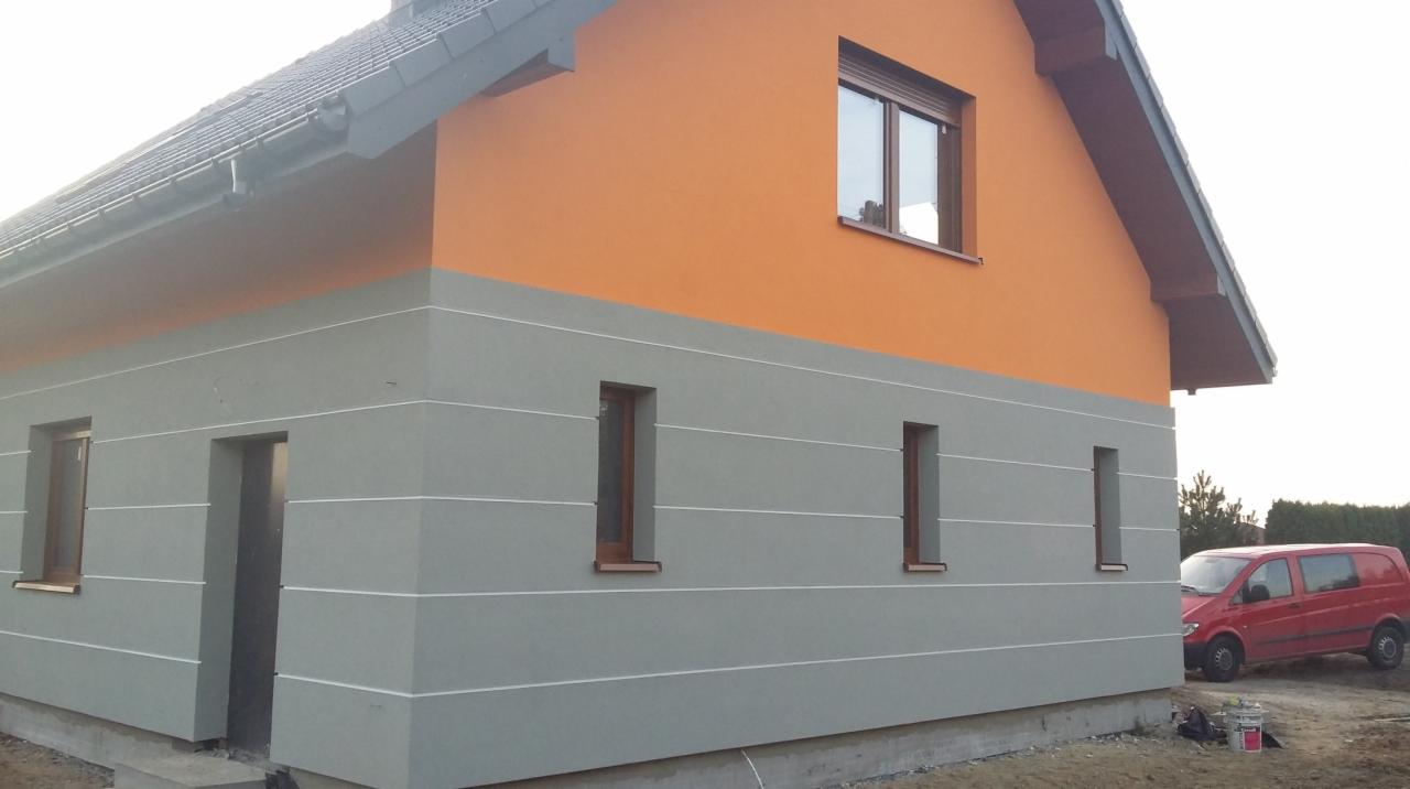 Dwukolorowa elewacja domu jednorodzinnego z poziomymi, białymi paskami. Widoczny fragment dachu, okna i czerwony samochód dostawczy w tle.