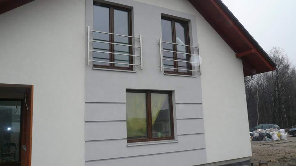 Dwukondygnacyjny dom z jasnoszarą elewacją, brązowymi oknami i metalowymi balkonami. Widoczne worki z materiałami budowlanymi przy ścianie.