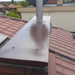 Błyszcząca, brązowa obróbka komina z blachy na dachu z dachówki ceramicznej, widoczny komin wentylacyjny i fragment instalacji fotowoltaicznej.