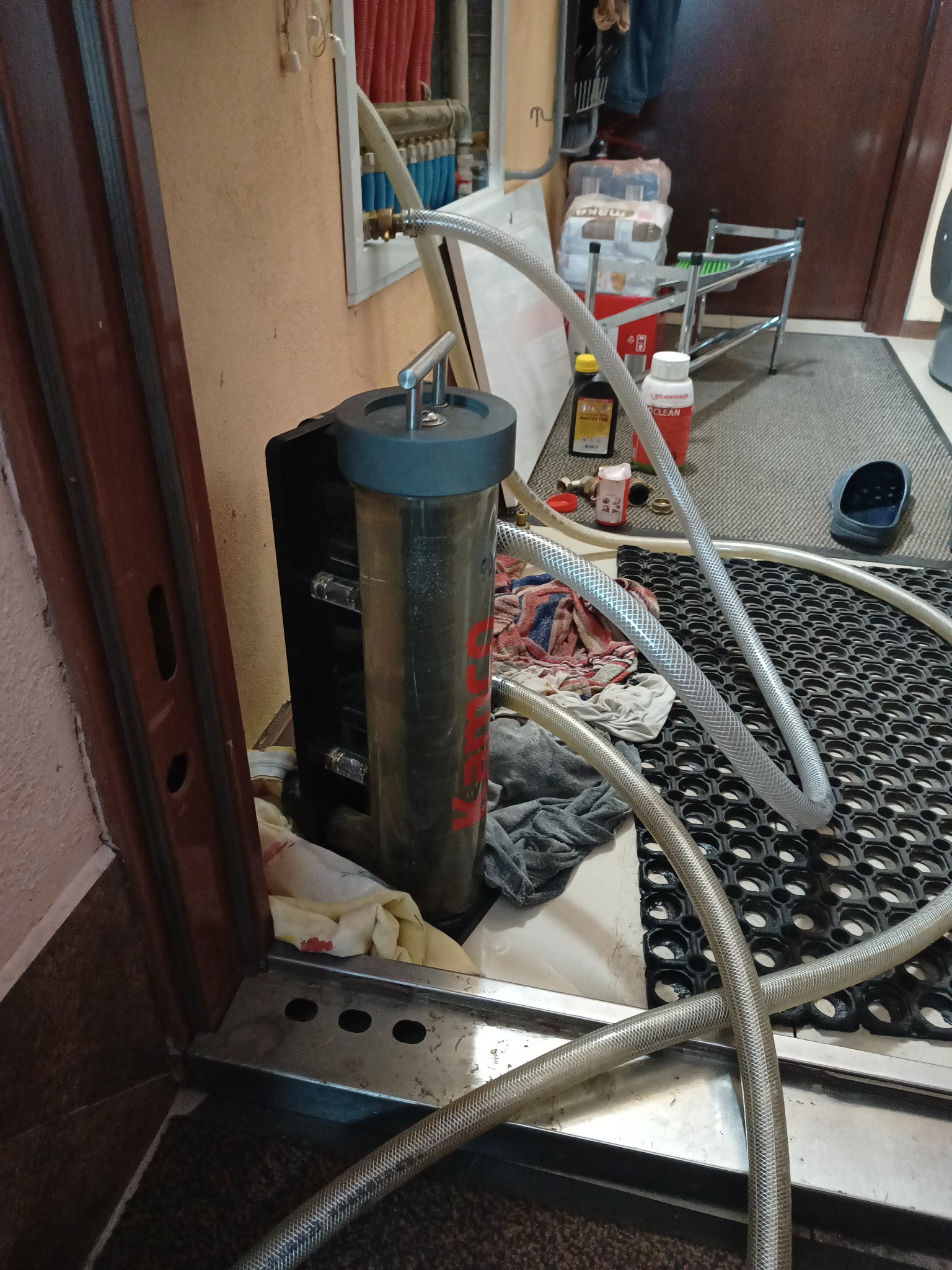 Filtr do instalacji grzewczej z podłączonymi wężami, stojący na podłodze w trakcie czyszczenia lub płukania instalacji. Widoczne środki czyszczące i akcesoria.