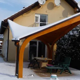 Nowoczesna drewniana konstrukcja z dachem pokrytym śniegiem, przylegająca do domu. Stół i krzesła pod spodem, zimowa sceneria.