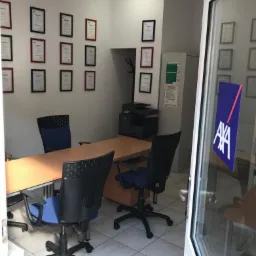 Wnętrze biura z biurkiem, krzesłami, ścianą z dyplomami w ramkach i logo AXA widocznym na szklanych drzwiach wejściowych.