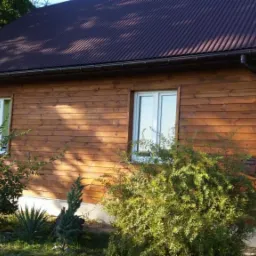 Dom jednorodzinny z drewnianą elewacją pomalowaną na brązowo, z brązowymi rynnami i brązowym dachem z blachy trapezowej, widoczny z poziomu gruntu w słoneczny dzień.