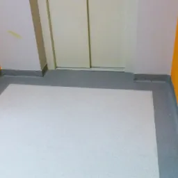 Biała wykładzina podłogowa z szarą listwą przypodłogową, pomarańczowe i zielone ściany, białe drzwi. Widok z góry, fragment wnętrza po remoncie.