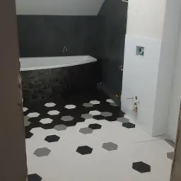 Wnętrze łazienki w trakcie remontu, z wzorem heksagonalnych płytek w odcieniach szarości i bieli na podłodze, czarna mozaika przy wannie, surowe ściany i instalacje w tle.