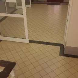 Beżowa podłoga wyłożona kwadratowymi płytkami z ciemnymi fugami, widoczny fragment białej balustrady schodów i otwarte białe drzwi wejściowe.