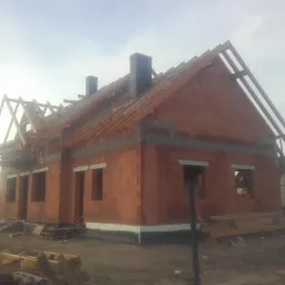 Budowa domu jednorodzinnego z czerwonej cegły, widoczne ściany, kominy i konstrukcja dachu w trakcie montażu, na dole stos drewna budowlanego.