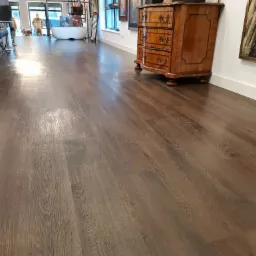 Nowoczesne wnętrze z ciemną, drewnianą podłogą ułożoną wzdłuż, oświetloną naturalnym światłem, z widoczną komodą z szufladami w stylu retro i elementami dekoracyjnymi w tle.