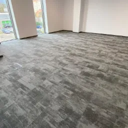 Szara, wzorzysta podłoga z płytek dywanowych w pustym, jasnym pomieszczeniu biurowym z dużymi oknami i widokiem na parking.