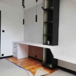 Białe biurko na wymiar z szufladami i drewnianym panelem, połączone z czarnym regałem z oświetleniem LED, w trakcie montażu, widoczne zabezpieczenia podłogi.