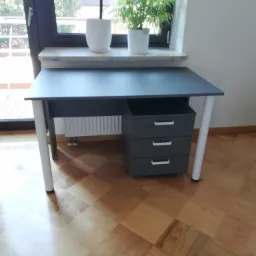 Szare biurko z trzema szufladami na kółkach, z białymi nogami, stojące na parkiecie, z dwoma doniczkami na parapecie w tle.