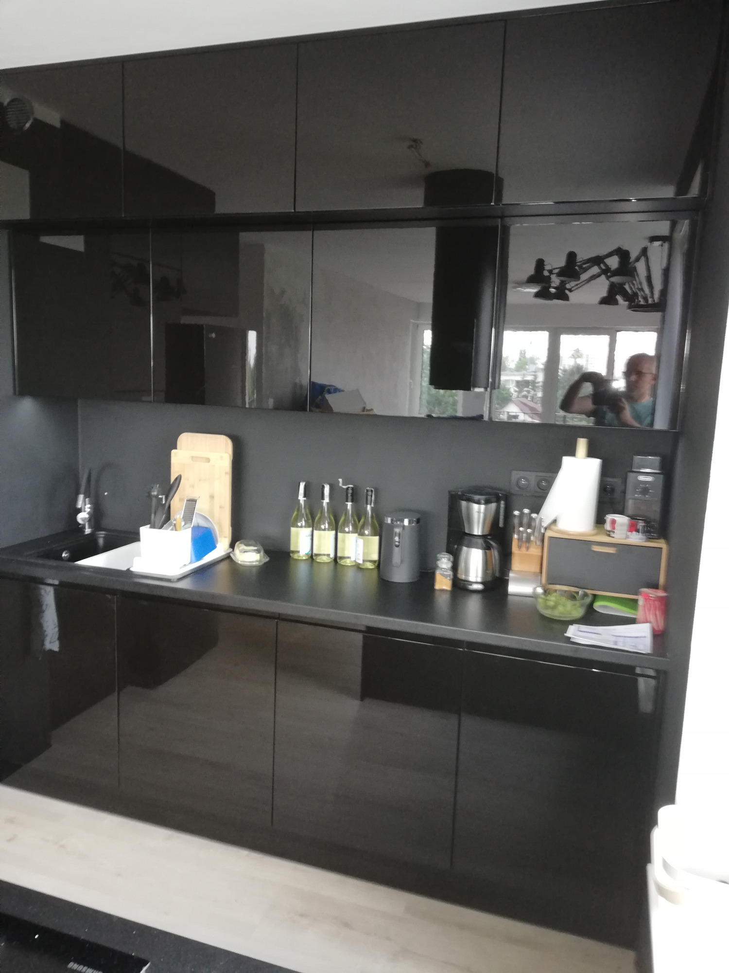 Czarna, błyszcząca zabudowa kuchenna z widocznym odbiciem osoby robiącej zdjęcie, na blacie kuchennym akcesoria, w zlewie naczynia.