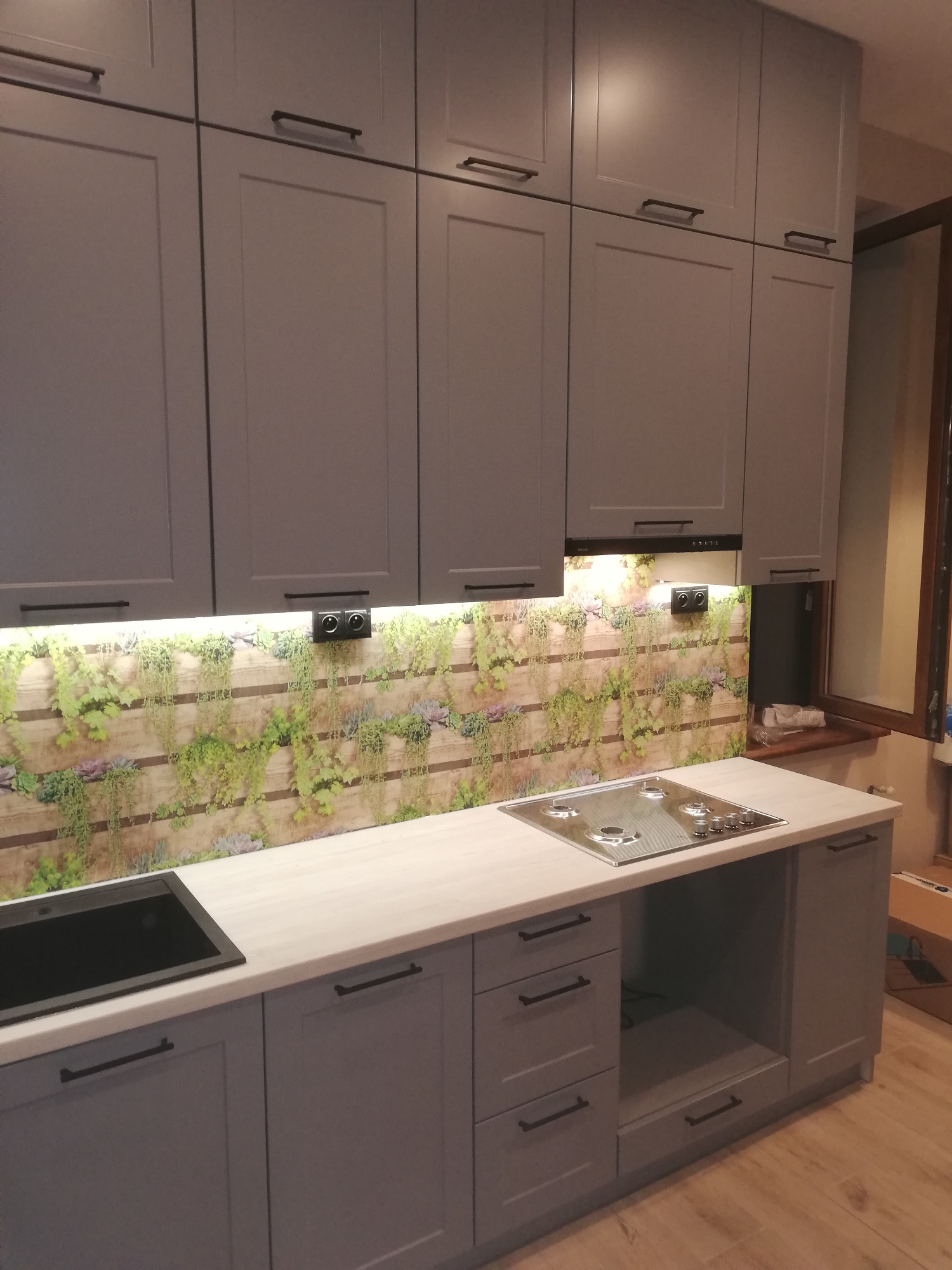 Szare szafki kuchenne z ciemnymi uchwytami, blat w kolorze jasnego drewna, ściana nad blatem z motywem roślinnym na drewnianym tle, płyta grzewcza i zlewozmywak.