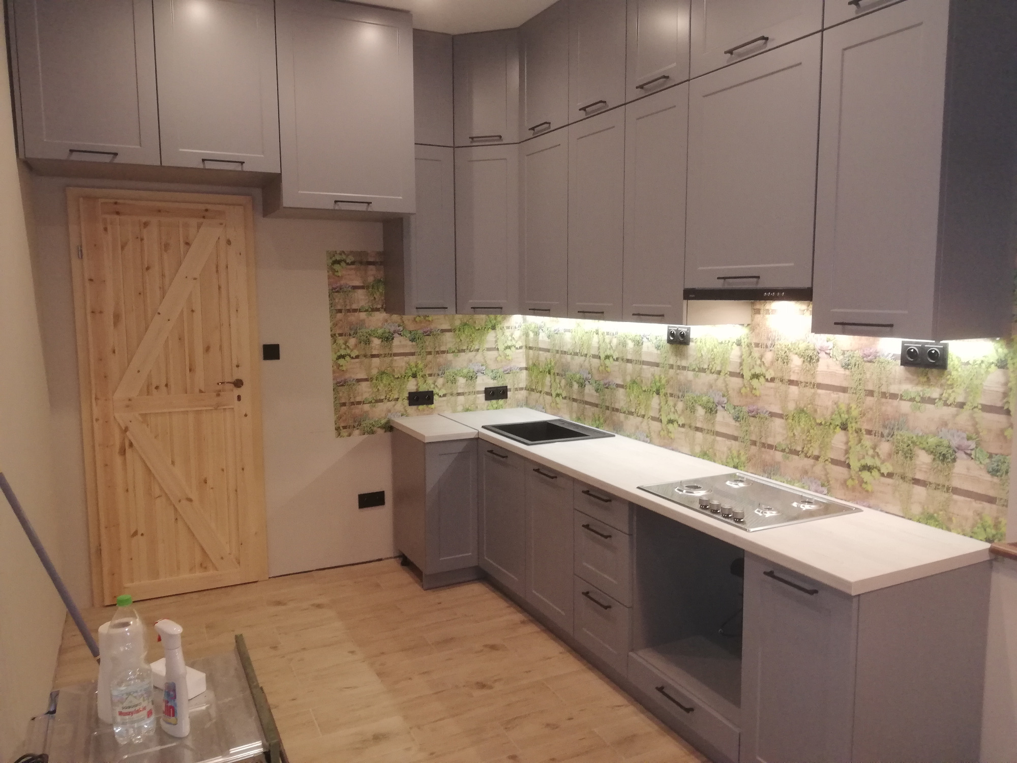 Szara kuchnia na wymiar z drewnianymi frontami szafek dolnych, blatem w jasnym odcieniu drewna i dekoracyjną tapetą imitującą roślinność na ścianie.