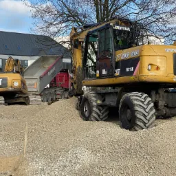 Żółty, kołowy koparko-ładowarka CAT M315D na placu budowy z wysypującą ziemię ciężarówką w tle. Widoczny budynek w tle i drzewa bez liści.