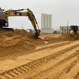 Żółty koparko-ładowarka CAT i wywrotka w trakcie prac ziemnych w Pile. W tle widoczne silosy cementowe i wyrównany teren z wyraźnymi śladami opon.