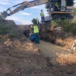 Jeziorowski i Syn - Prace ziemne: koparka CAT i pracownicy w żółtych kamizelkach przygotowują wykop, ubijając podłoże zagęszczarką. Widoczne ułożone kamienie i rów.