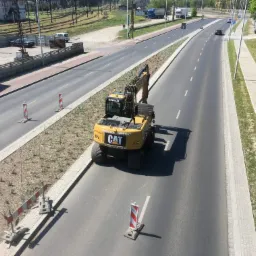 Żółta koparka CAT stoi na asfalcie w trakcie prac ziemnych przy drodze, widoczne słupki ostrzegawcze i świeżo nasadzona roślinność.