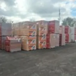 Sztaple materiałów budowlanych na placu, owinięte folią, widoczne opakowania z logo producenta, tło stanowią drzewa i pochmurne niebo.